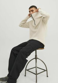 Beige hoog gesloten pullover met een ritssluiting en ribbeltextuur, gecombineerd met zwarte cargo-broeken met zakken, zittend op een metalen kruk.