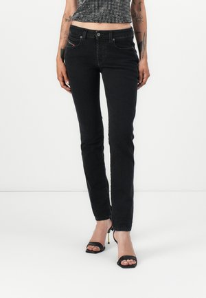 Femme portant un jean slim noir, des talons hauts noirs ouverts à l'avant et un top court à motifs noir et blanc, debout sur un fond blanc.