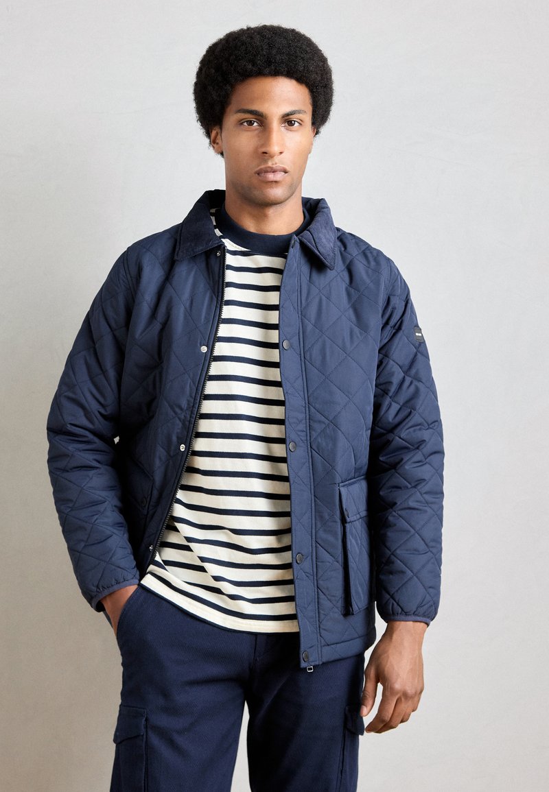 Mørkeblå quilted jakke med en kordfløjskrave, lynlås foran og frontlomme, båret over en stribet crew-neck t-shirt.