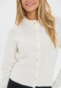 Usmiatu žena v bielej zapínacej kardigánovej bunde s rebrovaným lemom a dlhými rukávmi, s dlhými blond vlasmi prehodenými cez jedno plece.