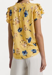 Gul blus med blommönster i blått och rosa, korta volangärmar och en samlad halsringning. Mjuk textur.