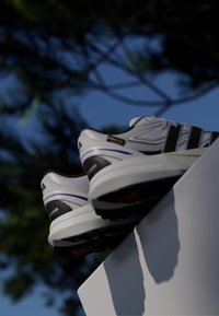 Bílé a černé atletické boty s tlustými podešvemi položeny na bílém okraji s rozostřeným pozadím stromu a modré oblohy.