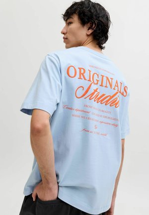 Hombre con cabello oscuro y rizado, que lleva una camiseta azul claro con el texto grande naranja "Originals Studio" en la espalda, con la mano en el bolsillo de los pantalones negros.