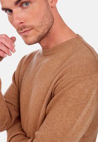 Homme avec une légère barbe et des yeux bleus, portant un pull en laine beige, posé avec la main près du visage contre un fond blanc uni.