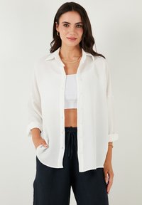 White, lightweight, button-up shirt with a textured pattern, rolled sleeve cuffs, worn over a white crop top paired with dark pants.