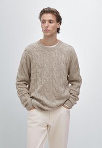 Pull beige en maille torsadée avec encolure ronde, motif texturé et poignets côtelés. Assorti à un pantalon clair, mains dans les poches.