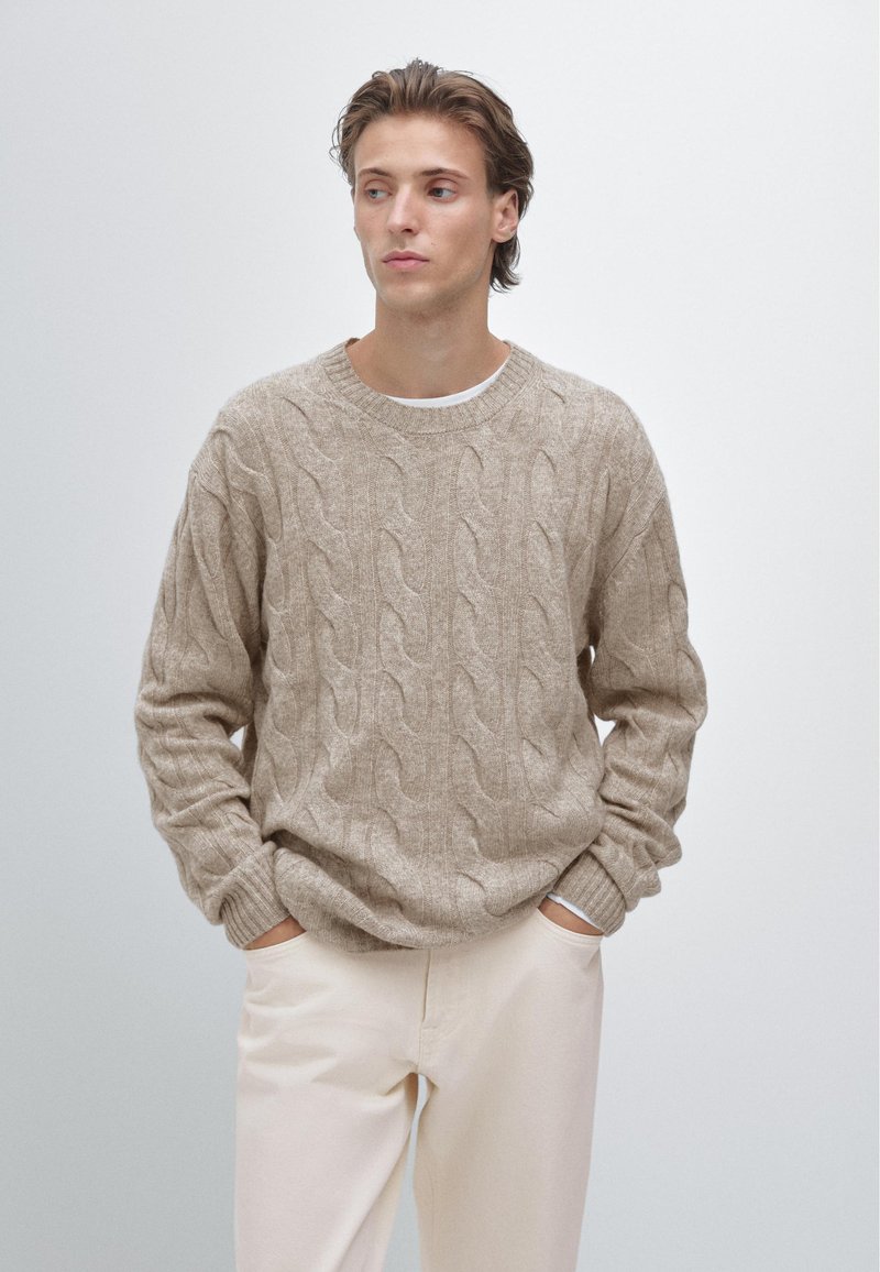 Pull beige en maille torsadée avec encolure ronde, motif texturé et poignets côtelés. Assorti à un pantalon clair, mains dans les poches.