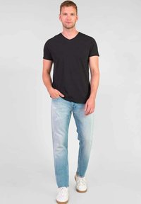 Zwarte V-hals t-shirt, lichtblauwe jeans met vervaagde details, en witte sneakers met lichtbruine zolen. Model staat tegen een effen achtergrond.