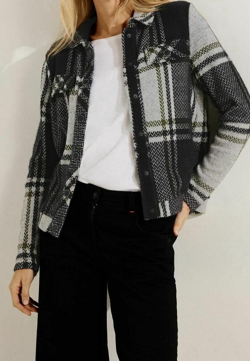 Femme portant un cardigan à carreaux noir et gris boutonné sur une chemise blanche, associé à un pantalon noir, debout avec une main dans la poche.