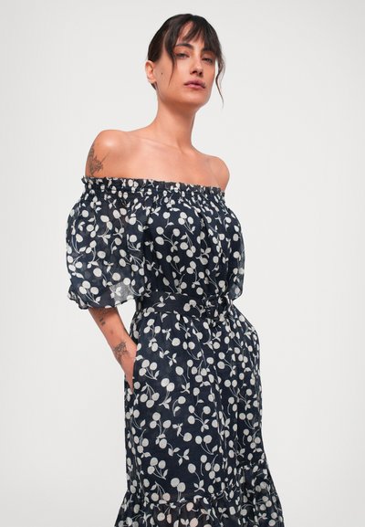 Frau mit Tattoos, die ein schulterfreies navy-blaues Kleid mit weißem Blumenmuster und Gürtel trägt, steht mit den Händen in den Taschen vor einem schlichten Hintergrund.