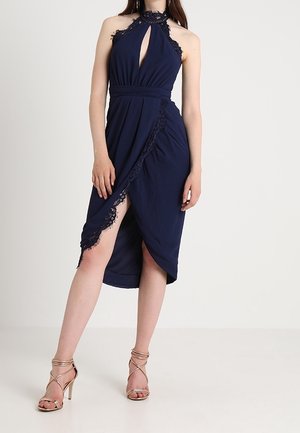 Vrouw draagt een marineblauwe mouwloze halterjurk met kanten rand en een hoge split, gecombineerd met gouden sandalen met hoge hak en bandjes.