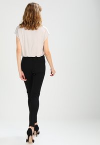Sorte tætsiddende bukser og en lys grå løs bluse, parret med sorte højhælede sko, set fra bagsiden mod en neutral baggrund.