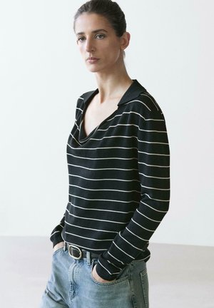 Kobieta z włosami związanymi do tyłu, ubrana w czarną bluzę z długim rękawem z białymi paskami i jasnoniebieskie dżinsy, stojąca z rękami w kieszeniach.