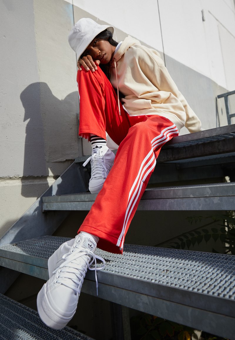 Zapatillas altas blancas, pantalones deportivos rojos con rayas blancas y una sudadera beige. Sentado en una escalera de metal en un entorno bien iluminado.