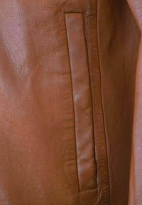 Braune Lederjacke mit einer glatten Textur, die über eine schlanke Seitentasche verfügt, die in das Design integriert ist. Subtile Strukturvariationen sind sichtbar.