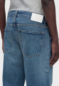 Gros plan d'une personne à la peau foncée portant un jean en denim bleu avec deux poches arrière et un patch en cuir blanc rectangulaire sur la ceinture.