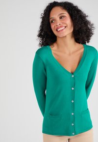 Cardigan verde con scollatura a V, chiusura a bottoni e due tasche frontali. Il materiale ha una texture liscia e dettagli a coste lungo i bordi.