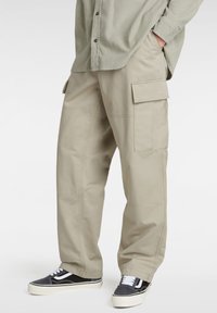 Beige cargo pants with side pockets, loose fit, and visible stitching details, paired with black and white sneakers.