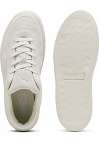 Białe zamszowe sneakersy Puma z białymi sznurówkami pokazane z góry obok ich teksturowanej podeszwy z logo Puma.