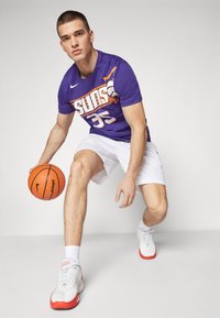 Paarse basketbaltricot met het "Suns" logo en nummer 35, gecombineerd met witte shorts en wit-rode basketbalschoenen, terwijl er een oranje basketbal wordt vastgehouden.