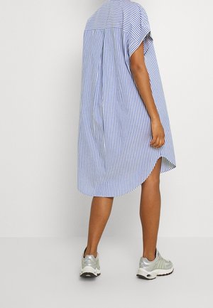 Mujer vestida con un vestido de manga corta a rayas azules y blancas con dobladillo curvo, llevando zapatillas deportivas grises y blancas sobre un fondo liso.
