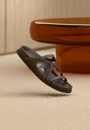 Brun læder slide sandal med krydsede remme og guld "Furla" branding, svævende over et beige tæppe nær en ravfarvet rund bordfod.
