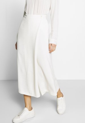 Mujer con blusa blanca de manga larga, pantalones blancos de talle alto y pierna ancha, y zapatillas blancas, de pie contra un fondo liso.