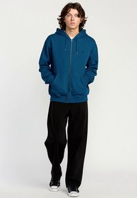 Sudadera con cremallera en color azul verdoso, con un gran bolsillo frontal, cordones ajustables y puños acanalados, combinada con pantalones negros de pierna ancha y zapatillas.