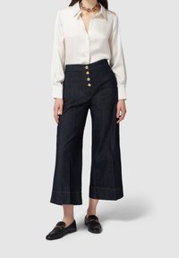 Bílá blůza s dlouhým rukávem v kombinaci s tmavě modrými kalhotami s vysokým pasem a širokými nohavicemi, které mají zlaté knoflíkové detaily a kontrastní švy. Černé mokasíny.