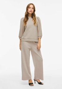 Felpa beige con maniche a sbuffo, abbinata a pantaloni a gamba larga coordinati. Texture liscia, design casual. Sandali neri con tacco alto completano il look.