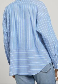 Blårandig skjorta med långa ärmar, med vita vertikala ränder, en lös passform och en rundad fåll baktill. Bomullstextur.