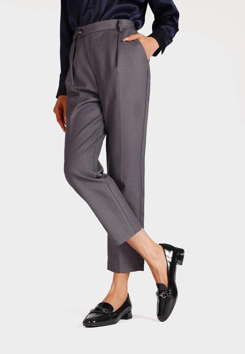 Pantaloni grigi su misura con vita alta e pieghe, abbinati a mocassini neri lucidi con un accento dorato. Tessuto dalla texture liscia.