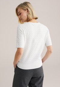 Biały sweter z krótkim rękawem z fakturowanym trójkątnym wzorem, ściągaczami na mankietach i dole, zestawiony z szarymi spodniami w paski.