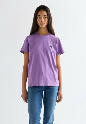 Młoda kobieta o długich ciemnych włosach, ubrana w fioletowy t-shirt i niebieskie dżinsy, stojąca na tle jednolitego białego tła.