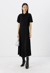 Schwarzes, kurzärmeliges Hemdkleid mit fließendem Design und einer Länge bis zur Mitte der Waden. Kombiniert mit hohen schwarzen Stiefeln und einer kleinen silbernen Tasche.