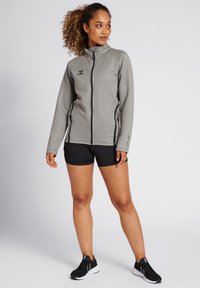 Grå fleecejakke med høj krave, lynlås foran og sorte detaljer. Bærer den over sorte shorts og sorte sneakers.