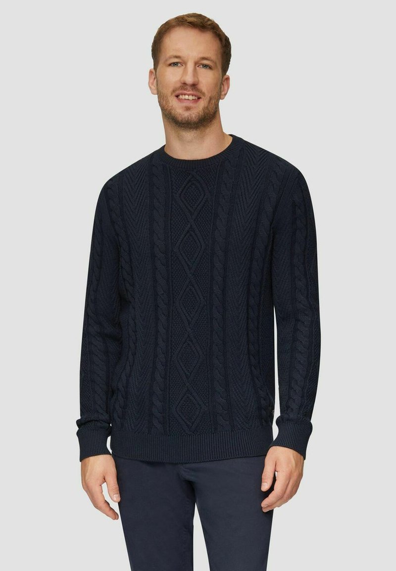 Ein Mann lächelt, trägt einen dunkelblauen Kabelstrickpullover und passende dunkle Hosen und steht vor einem schlichten hellen Hintergrund.