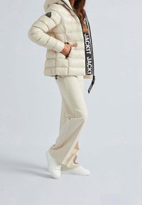 Polstret beige jakke med mørke remdetaljer, kombineret med cremefarvede bukser og hvide sneakers, med lynlåslommer og hætte.