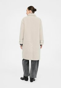 Donna con i capelli raccolti in un chignon, che indossa un lungo cappotto di pile beige, jeans larghi grigi e stivali neri alla caviglia, in piedi con la schiena rivolta.