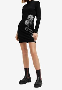 Svart långärmad klänning med vit blommig broderi på sidan, tillverkad av elastiskt material. Kombineras med svarta stövlar med röda accenter.