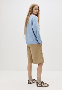 Hellblaues, übergroßes Sweatshirt, beige Shorts, gepunktete Schuhe. Lange, lockige Haare, stehend mit dem Rücken zu einem schlichten weißen Hintergrund.