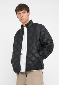Giacca a vento trapuntata nera con colletto alto, dotata di chiusura con zip e polsini elasticizzati, indossata sopra una camicia bianca e pantaloni beige.