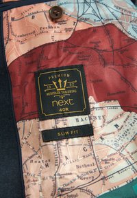 Niebieskie wnętrze z tkaniny z wzorem mapy w beżowych i czerwonych odcieniach. Etykiety zawierają napisy "PREMIUM HERITAGE TAILORING" oraz "SLIM FIT."