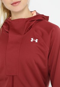 Giacca cappuccio bordeaux con accenti rosa chiaro, dotata di chiusura a zip e logo Under Armour bianco sul petto. Tessuto liscio e leggero.