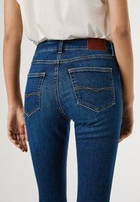 Jeans de mezclilla azul oscuro con un diseño ajustado, que cuentan con dos bolsillos traseros con costuras amarillas y una etiqueta de cinturón de cuero marrón.