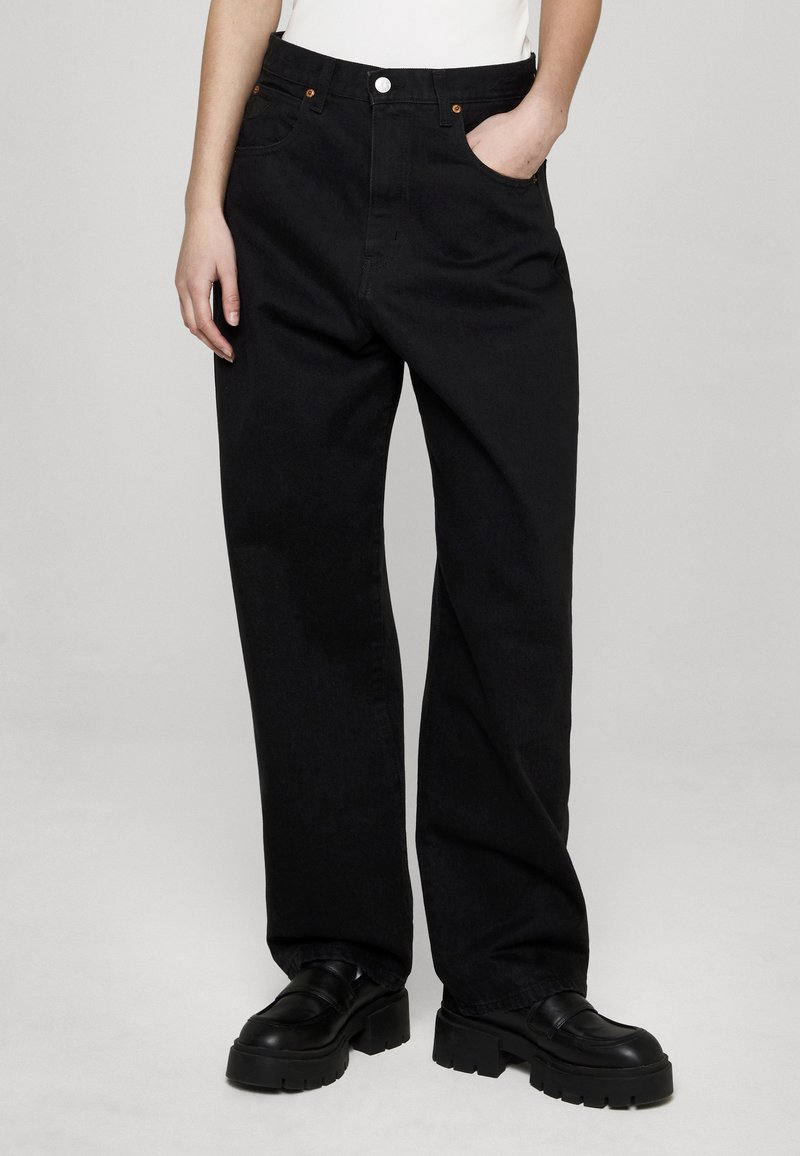 Vaqueros negros de tiro alto y pierna recta hechos de mezclilla, con cinco bolsillos, remaches de cobre y una textura sutil. Combinados con zapatos de plataforma negros.