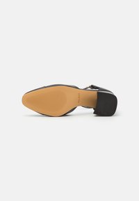Zwarte high heel schoen met een spitse neus, voorzien van een glad bovenwerk, stevige blokhak en een getextureerde tan rubberen zool.