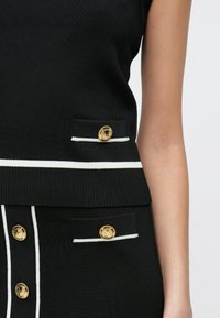 Μαύρο πλεκτό δίχρωμο σύνολο που περιλαμβάνει αμάνικο τοπ με λευκές ρίγες και διακοσμητικά κουμπιά σε χρυσό χρώμα, καθώς και φούστα με παρόμοιες λεπτομέρειες κουμπιών.