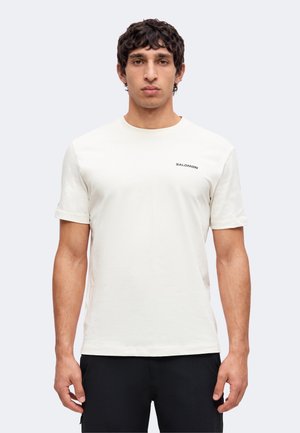 Jeune homme aux cheveux bouclés foncés portant un t-shirt blanc uni à manches courtes Salomon et un pantalon noir, debout devant un fond clair.