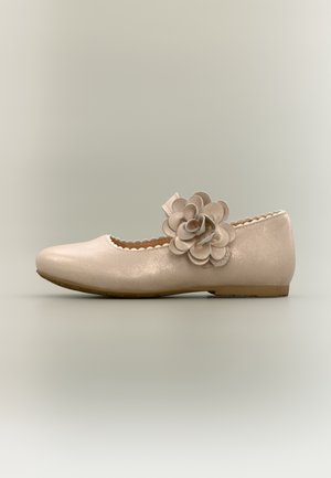 Beige flad sko med bølget pynt og stor dekorativ blomst på rem over vristen, vist på en neutral baggrund.